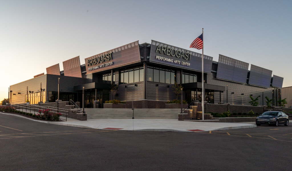 Exterior view of the Arbogast Performing Arts Center, featuring modern architecture, large windows, an American flag, and a surrounding parking lot at sunset.