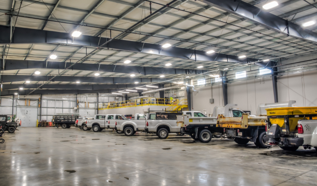 A large, well-lit industrial warehouse with numerous work trucks and heavy-duty vehicles parked inside. The concrete floor is clean and the ceiling has bright overhead lights.