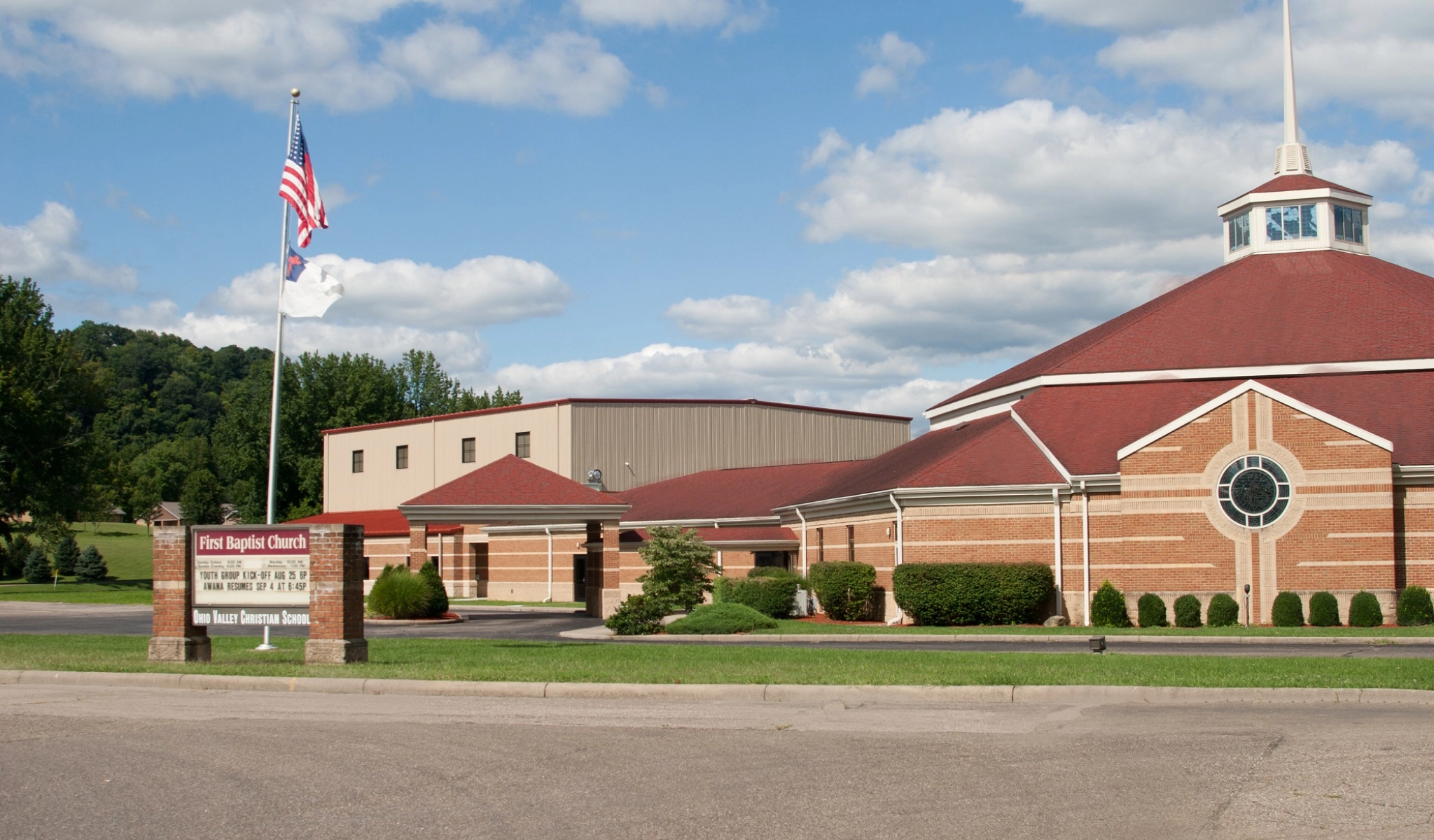 A brick building with a red roof and steeple, known as the First Baptist Church, is identified by a sign by the road. An American flag and another flag fly on a pole. Green trees and bushes surround the property.