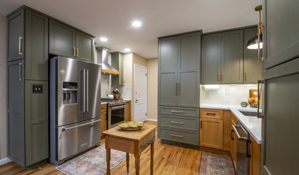 A modern kitchen with green and wood-tone cabinets, stainless steel appliances, a small wooden island, and hardwood flooring.