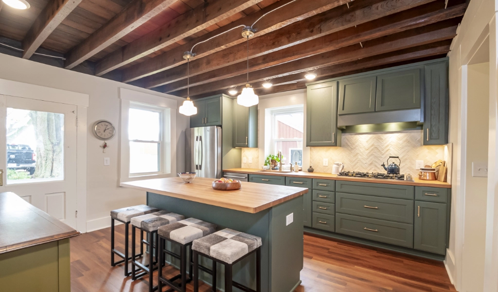 A modern kitchen features green cabinetry, a wooden countertop island with three stools, stainless steel appliances, pendant lights, and an exposed beam ceiling.