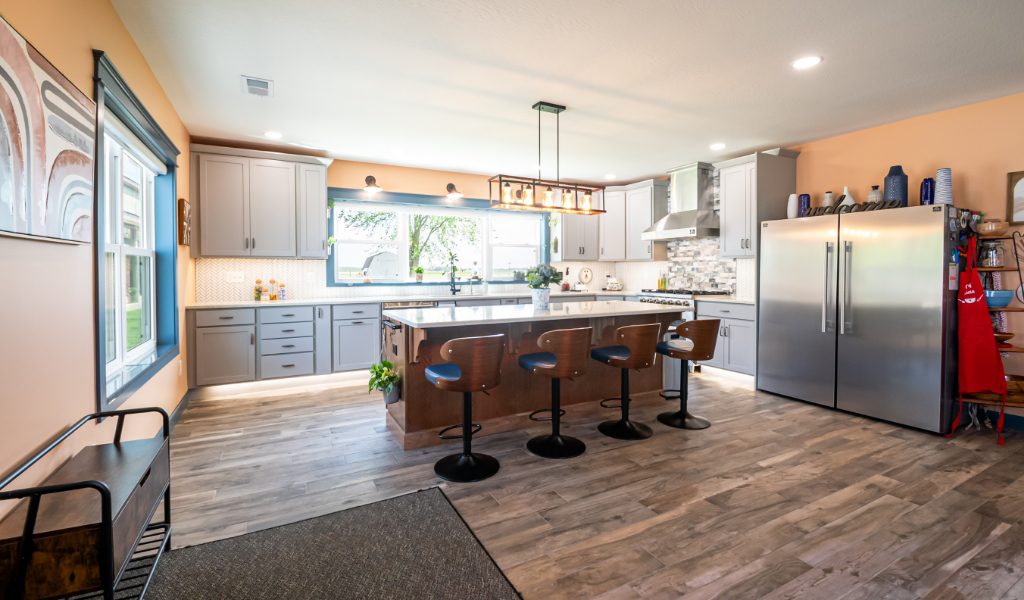 Modern kitchen with gray cabinets, a large island with four barstools, stainless steel appliances, wood flooring, and large windows letting in natural light—an award-winning home addition.