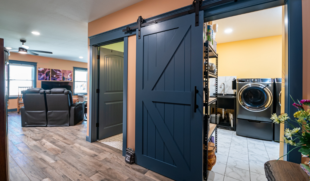 Home interior featuring an award-winning home addition: a blue sliding barn door reveals a laundry room with washer and shelving, next to a living area with recliners and ceiling fan.