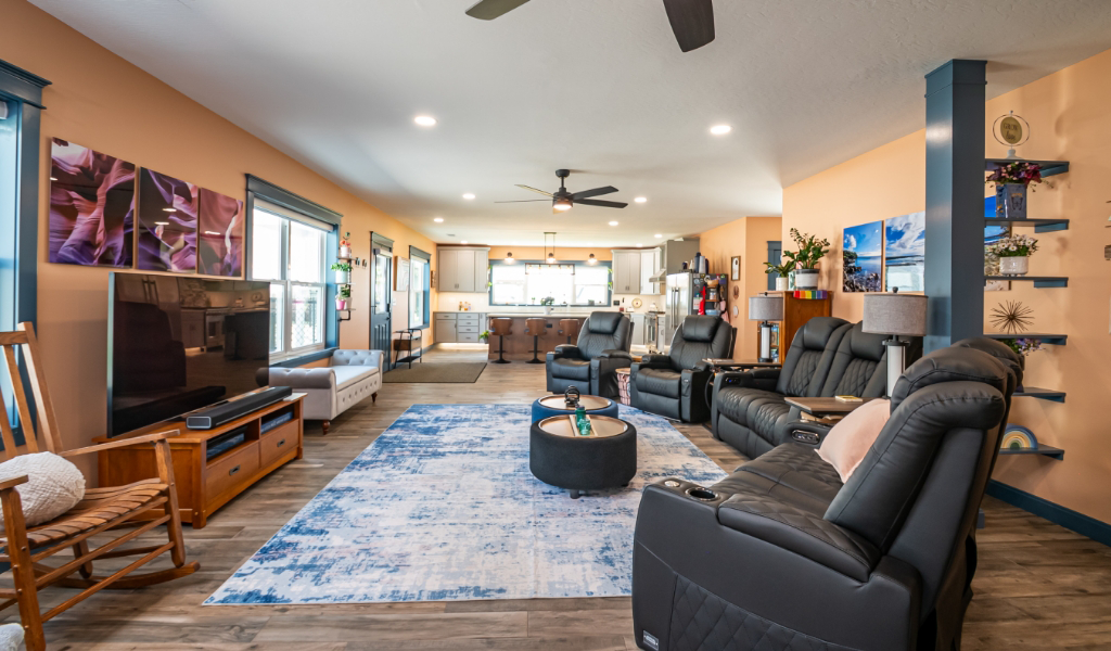 Spacious living room with black recliners, blue and white rug, wall art, large windows, and an open view into the kitchen area—all part of an award-winning home addition.