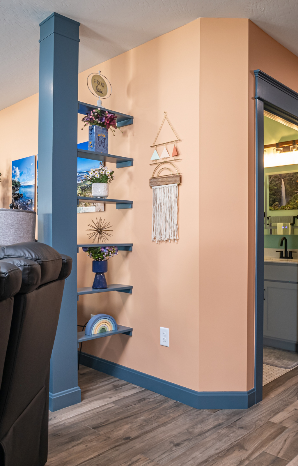 A corner wall with floating shelves displaying decor items, macrame wall hanging, and a view into an award-winning home addition featuring a bathroom with light teal cabinetry.