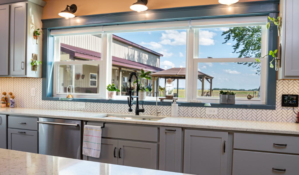 A modern kitchen with gray cabinets, white countertops, and a large window overlooking a yard with a gazebo and barn under a blue sky—part of an award-winning home addition.