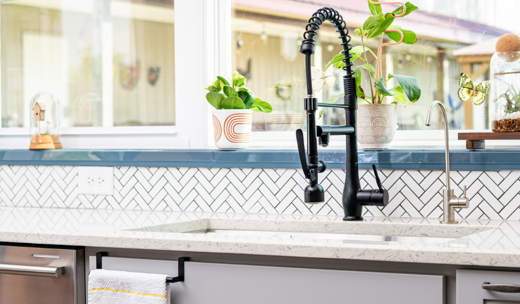Modern kitchen sink area with a black pull-down faucet, small plants on the windowsill, a towel hanging below, and herringbone tile backsplash—part of an award-winning home addition.