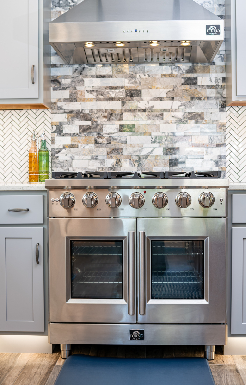 A stainless steel double oven with six stovetop burners is set against a tile backsplash, flanked by light gray cabinets and two bottles of oil in this award-winning home addition.