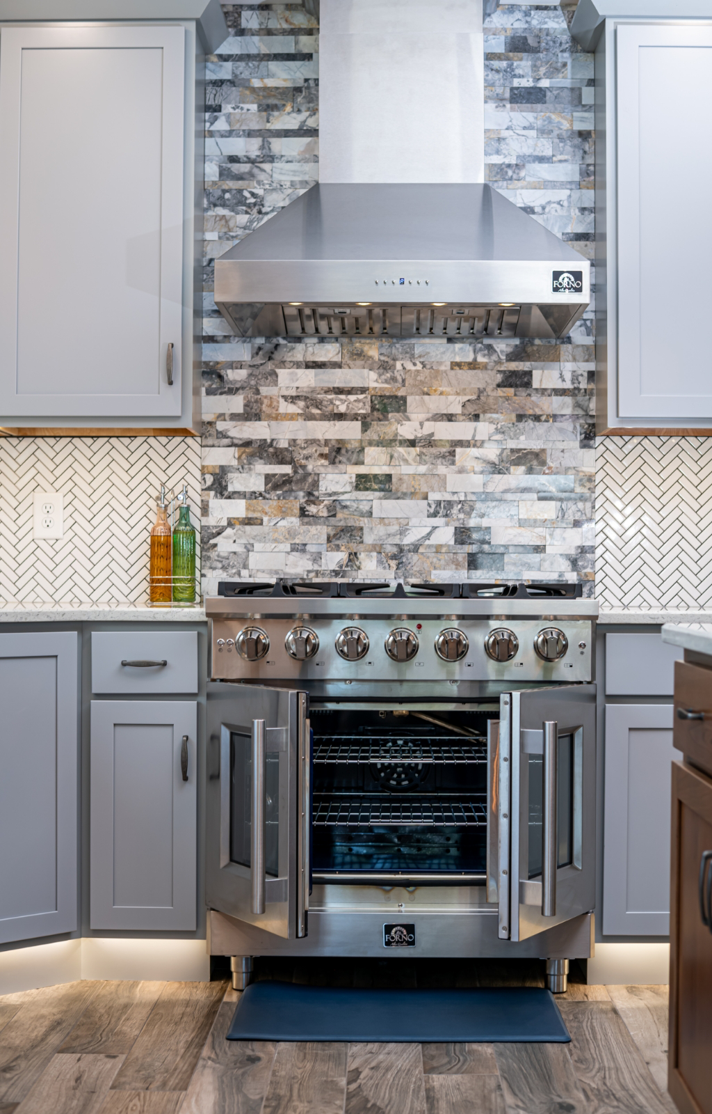 A modern kitchen in this award-winning home addition features a stainless steel oven with double doors open, gray cabinets, a tile backsplash, and a range hood above.