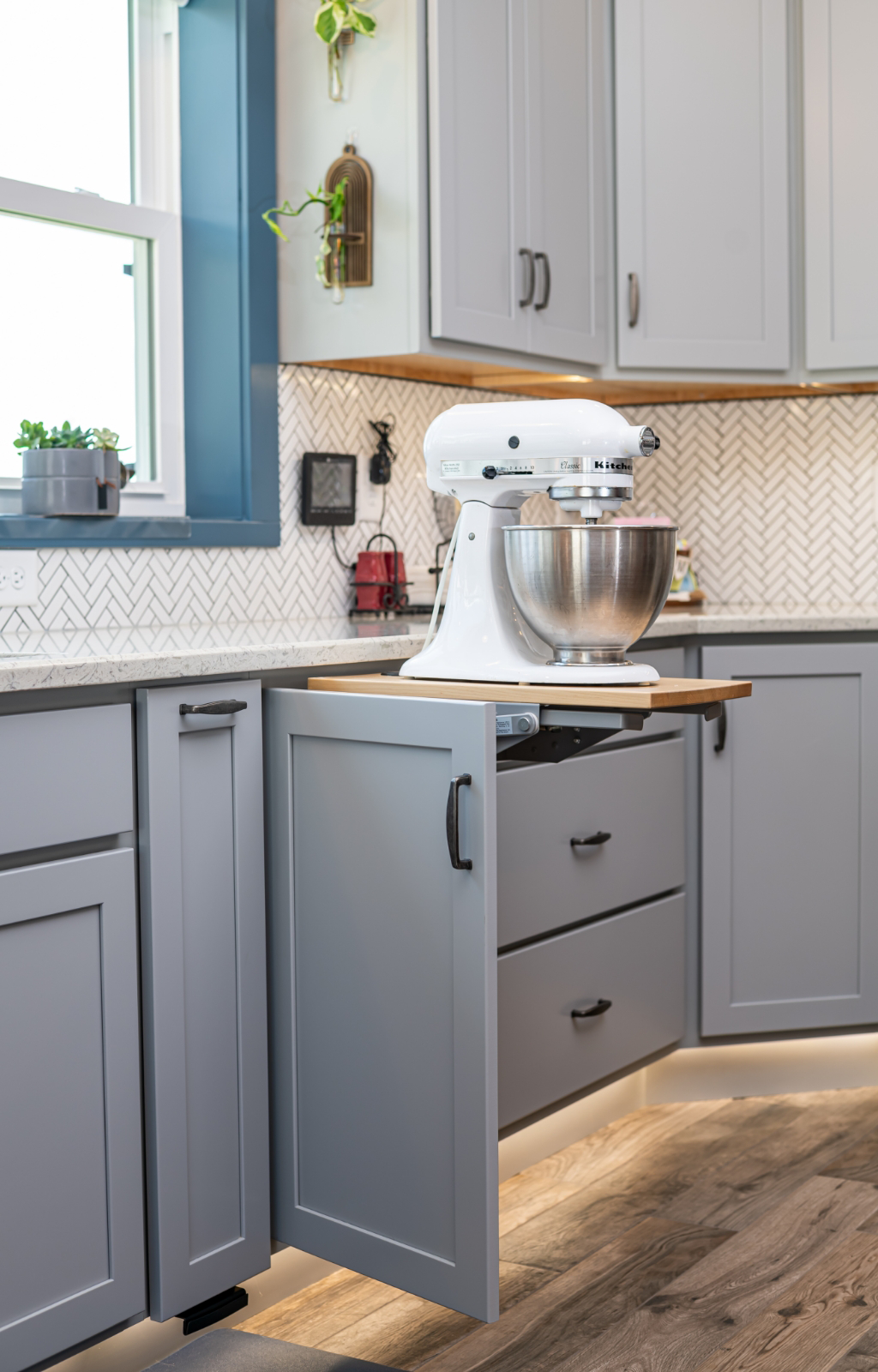 A stand mixer is placed on a pull-out cabinet shelf in a modern kitchen with gray cabinets and a chevron-patterned backsplash, part of an award-winning home addition.