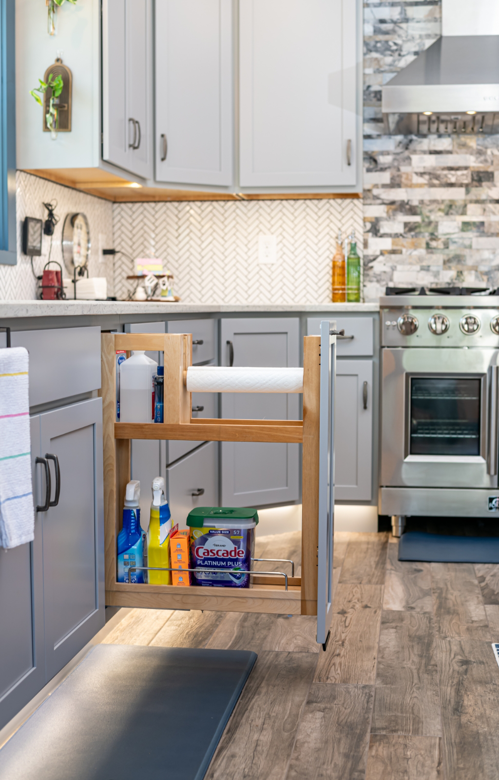 An award-winning home addition, this kitchen with gray cabinets features a pull-out drawer open, displaying cleaning supplies, a paper towel roll, and dishwashing detergent.