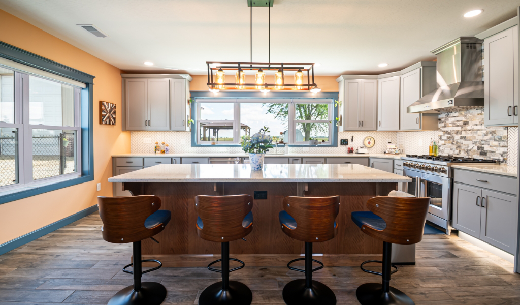 Modern kitchen with a large island, four wooden barstools, stainless steel appliances, and gray cabinets. Pendant lighting and large windows fill this award-winning home addition with natural light.