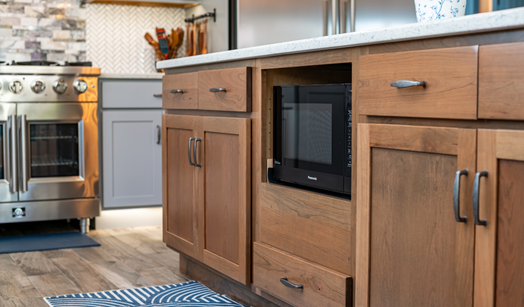 A built-in microwave is installed in a wooden kitchen cabinet next to a stove, with drawers and a rug visible on a wood floor—part of an award-winning home addition that blends function with style.