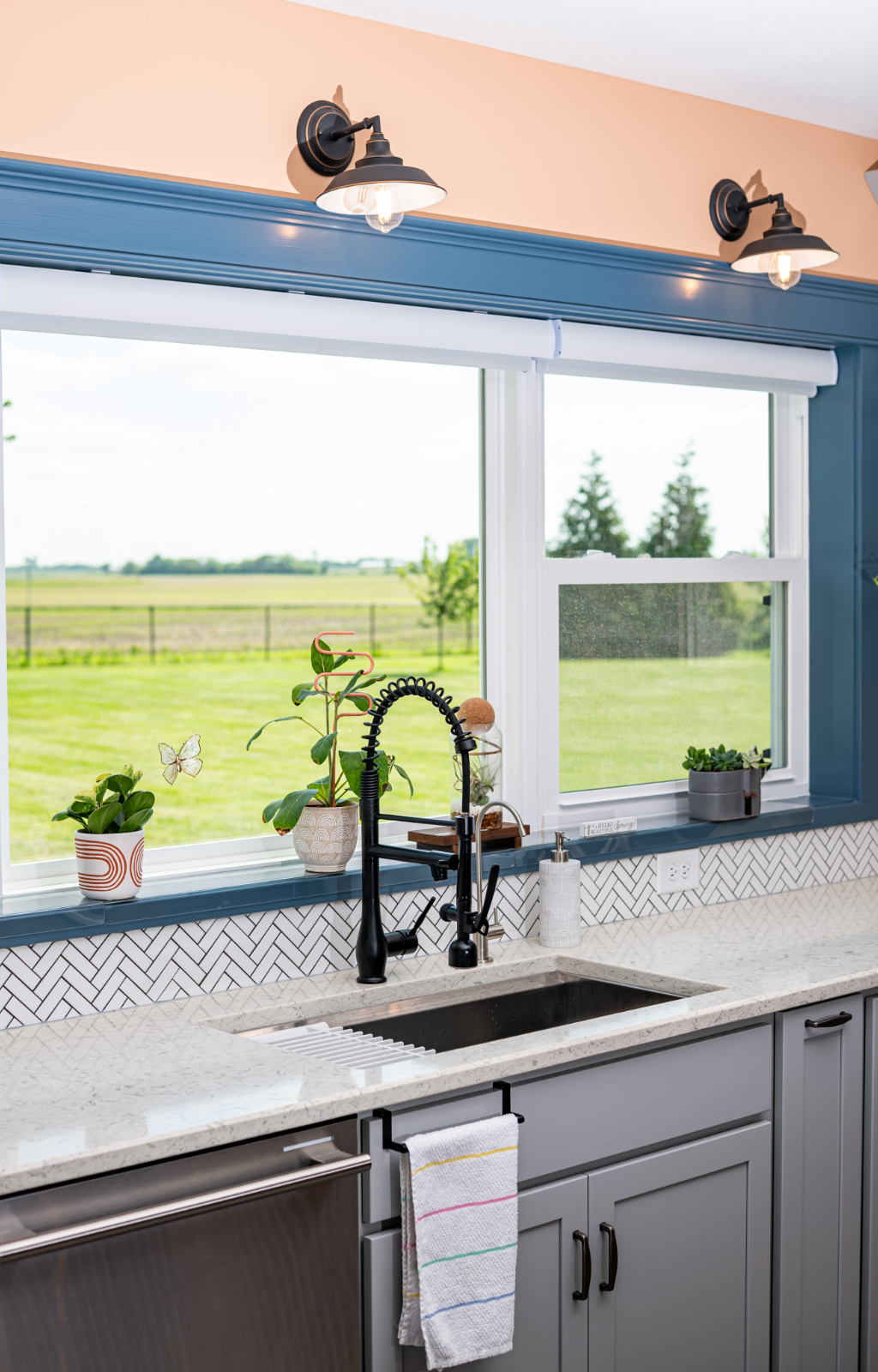 Bright kitchen sink area with a matte black faucet, grey cabinets, a white herringbone tile backsplash, potted plants, and large windows overlooking a green outdoor landscape in this award-winning home addition.
