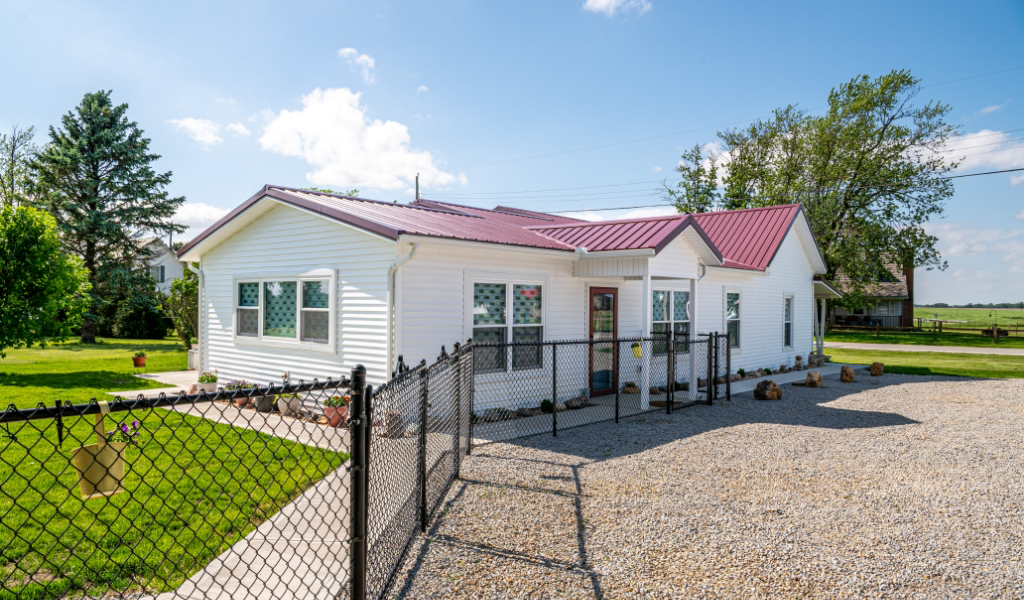 Single-story white house with a red roof, fenced yard, gravel driveway, and green lawn under a blue sky with scattered clouds—featuring an award-winning home addition for extra charm and space.