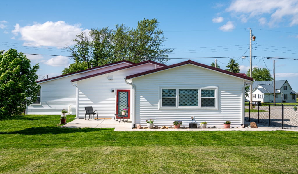 Single-story white house with a red roof, front patio seating, potted plants, and a grassy lawn under a partly cloudy sky—featuring an award-winning home addition for enhanced comfort and style.