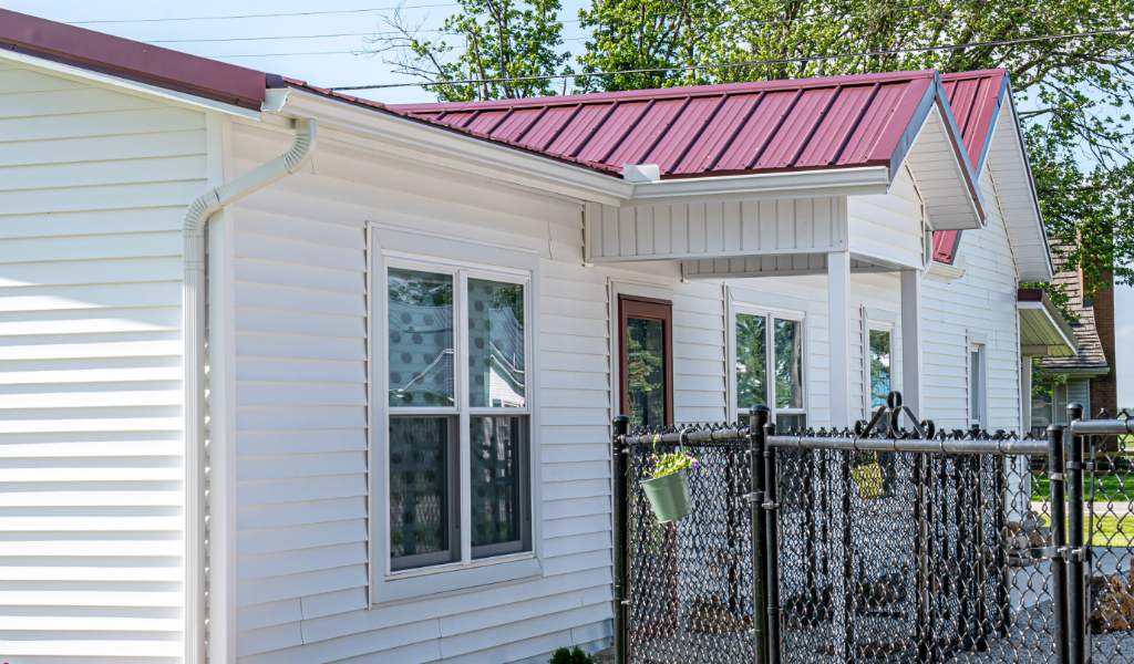 A white house with a red metal roof, white siding, several windows, and a chain-link fence in the front yard features an award-winning home addition.