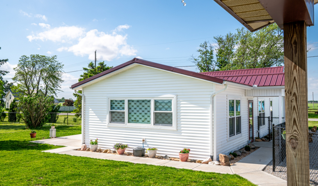 Single-story white house with a red metal roof, three windows, and several potted plants along the concrete sidewalk. This award-winning home addition sits on a grassy yard with trees in the background.