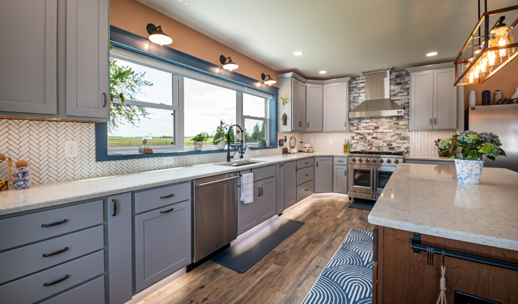 Spacious modern kitchen with gray cabinets, stainless steel appliances, large windows, and patterned backsplash. Part of an award-winning home addition, a potted plant sits on the island countertop.