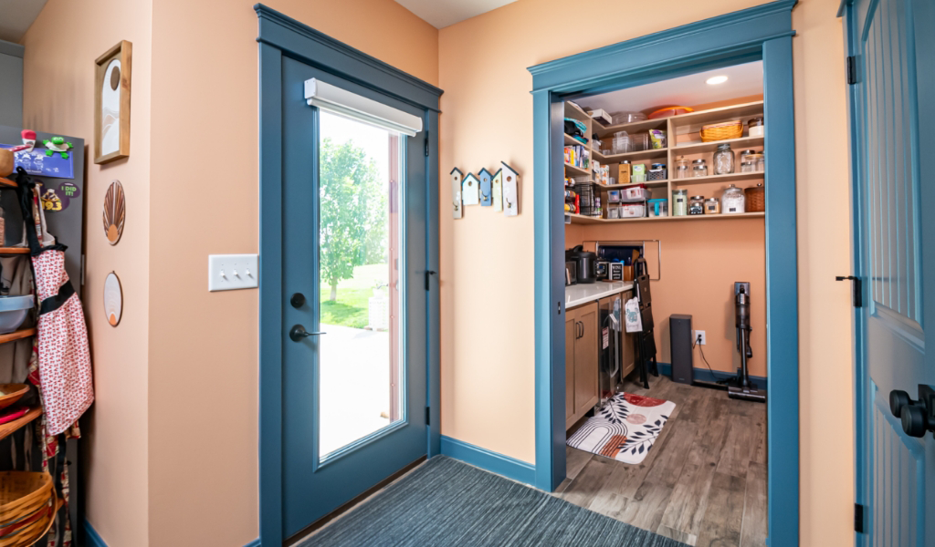 A mudroom with peach walls, blue trim, and a glass door to the outside is part of this award-winning home addition, which also features an adjacent organized pantry with shelves and small appliances.
