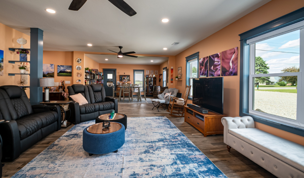 A spacious living room with black recliners, a blue and white rug, a large TV, colorful wall art, and a window view. This award-winning home addition seamlessly connects to a stylish dining area in the back.