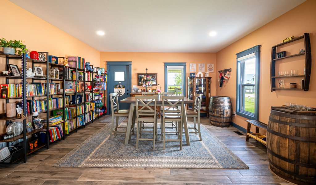 A cozy, award-winning home addition features orange walls, bookshelves, a central wooden table with chairs, two barrels, a rug, and windows letting in natural light.