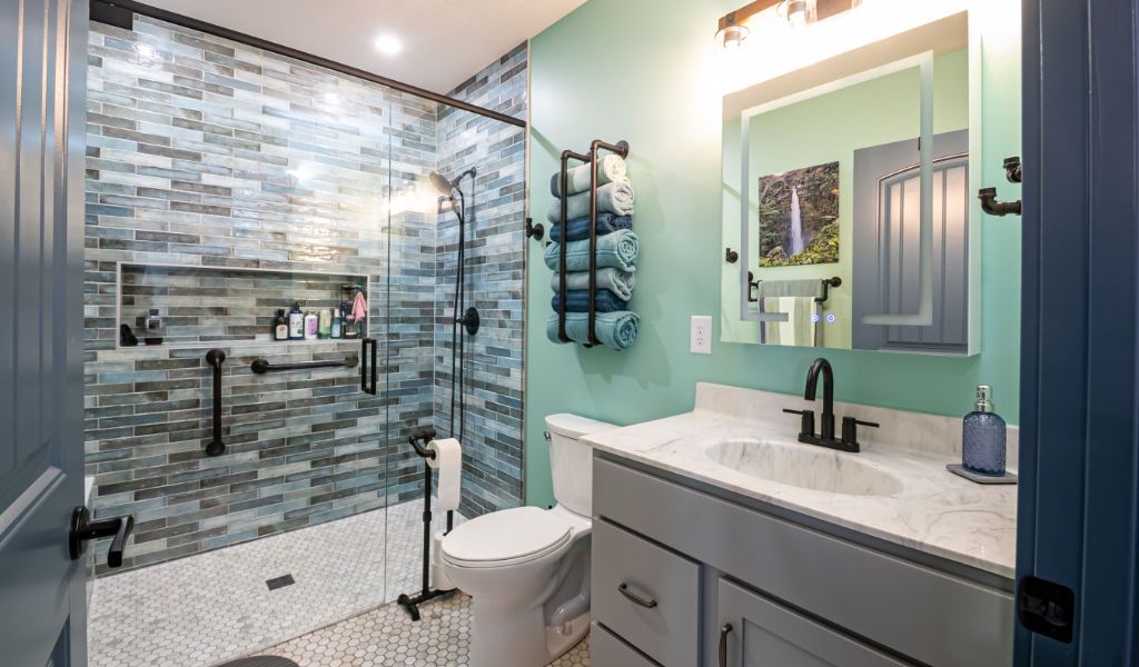 Modern bathroom with a glass-enclosed shower, folded towels on a wall rack, a toilet, and a vanity with sink and mirror under wall lighting on a light green wall in this award-winning home addition.