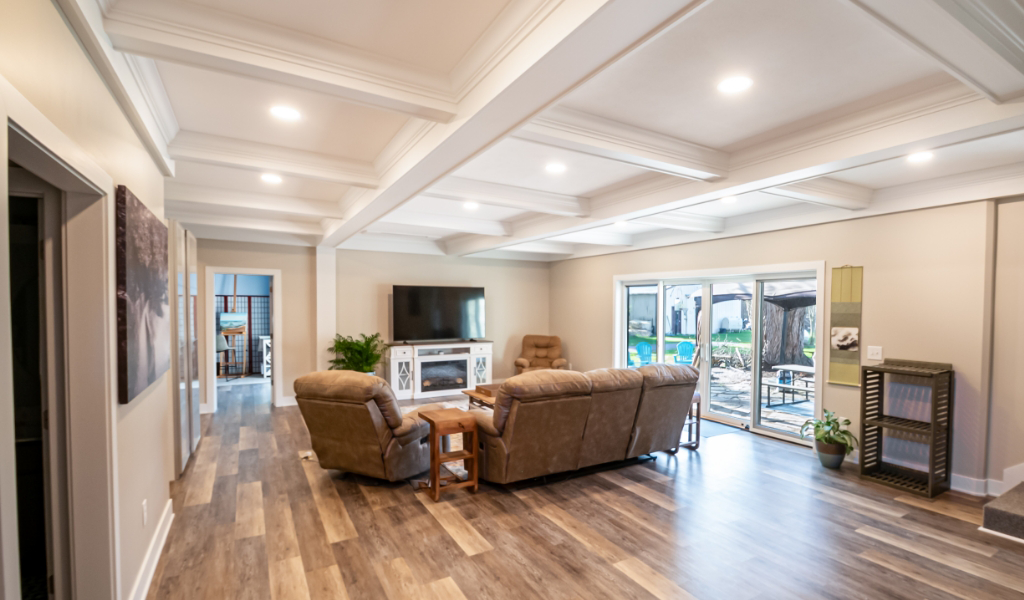 Bright living room with coffered ceiling, wood flooring, brown recliner sofas, a TV, and large glass doors opening to a patio and backyard—part of an award-winning home remodel & additions project.