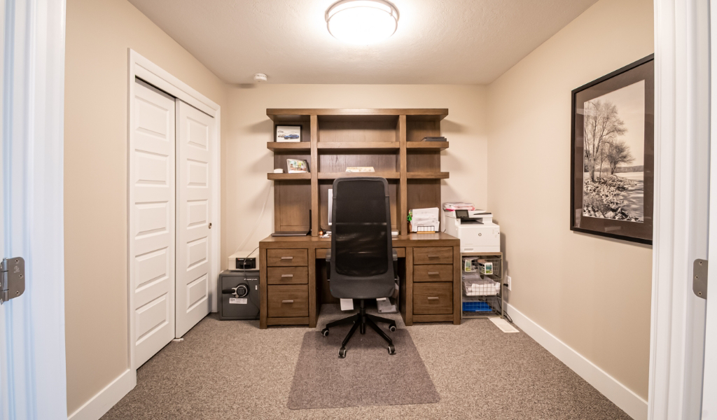 A home office from an award-winning home remodel & additions project, featuring a wooden desk, office chair, shelves, computer, printer, safe, and a framed landscape photo. The space boasts neutral walls and a carpeted floor.