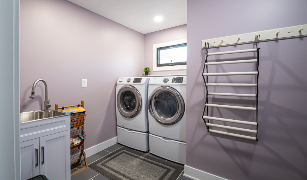 A modern laundry room, part of an award-winning home remodel & additions, features lavender walls, washer and dryer, utility sink, drying rack, window, and basket organizer for ultimate functionality and style.