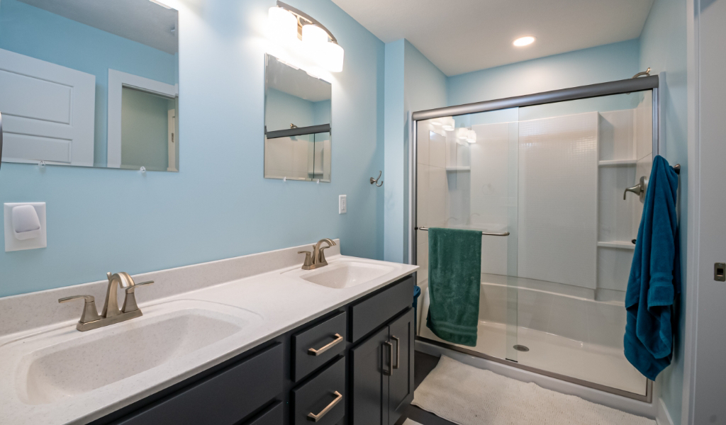 Modern bathroom with double sink vanity, two mirrors, and a glass-enclosed shower. Towels hang on the shower door, and the walls are painted light blue—an example of award-winning home remodel & additions.