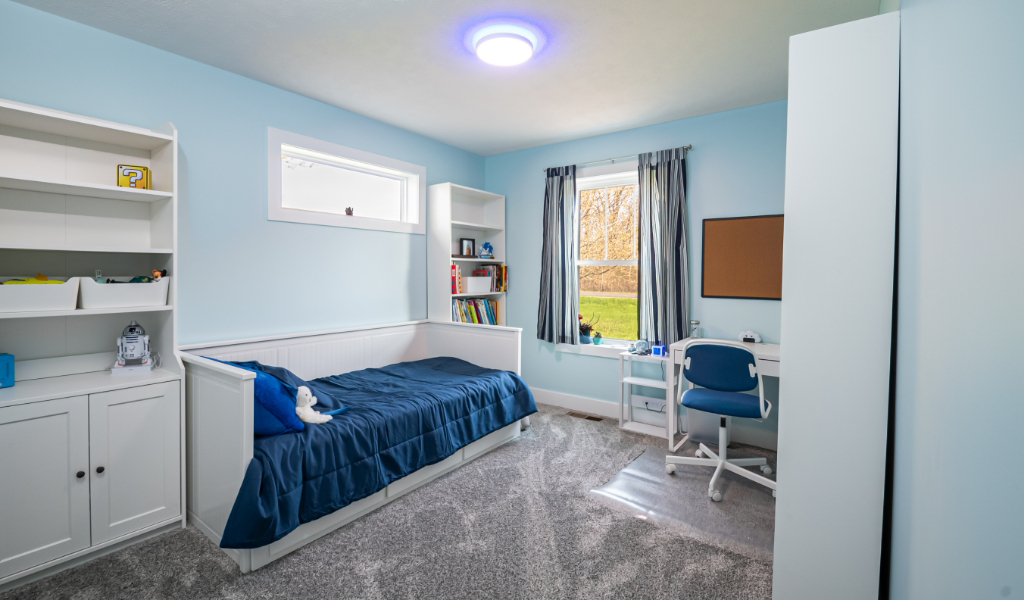 A tidy, modern bedroom with light blue walls, a single bed with blue bedding, a desk and chair by the window, shelves, and a carpeted floor—part of an award-winning home remodel & additions project.