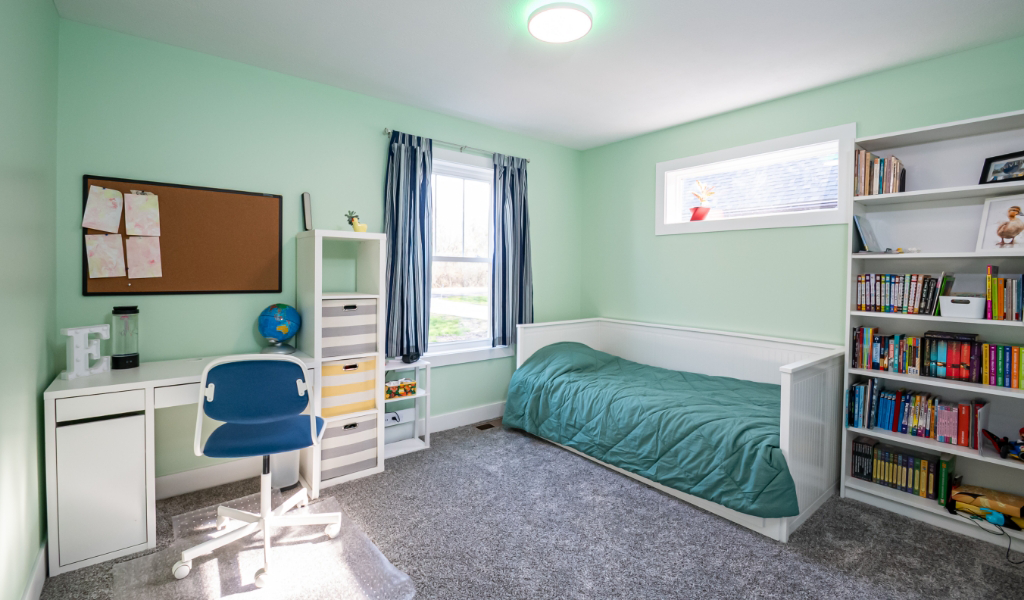 A tidy bedroom with a single bed, bookshelf, desk with chair, corkboard, and window with blue curtains; walls are painted light green and floor is carpeted gray—an example of award-winning home remodel & additions.