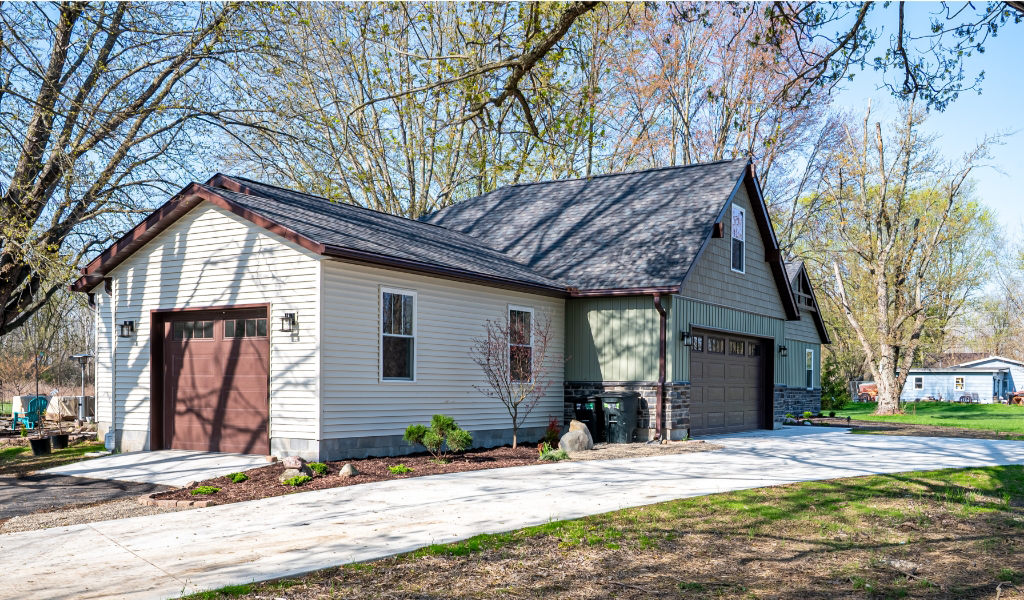 An award-winning home remodel features this modern detached garage with beige siding, a brown garage door, and gray stone accents, situated on a concrete driveway surrounded by trees and lawn.
