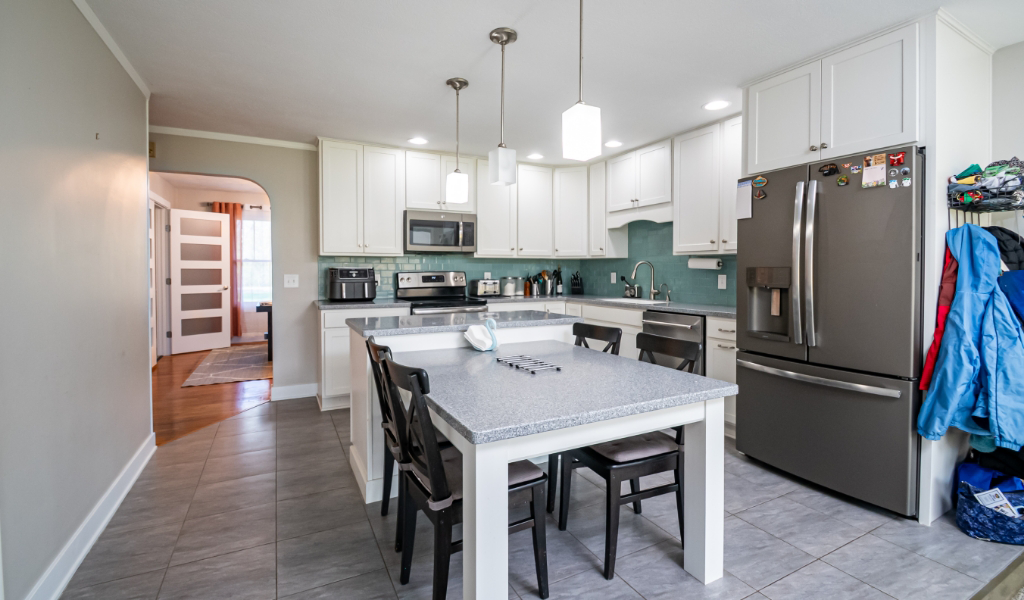 Modern kitchen from an Award-Winning Home Remodel & Additions, featuring white cabinets, stainless steel appliances, a gray island with chairs, and jackets hanging on hooks near the refrigerator.