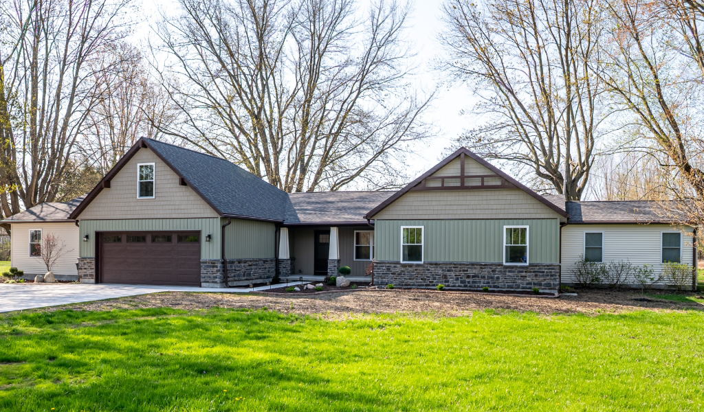 Award-Winning Home Remodel & Additions: Single-story house with a two-car garage, stone and siding exterior, surrounded by a green lawn and leafless trees in the background.