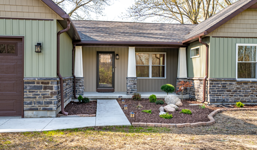 Single-story house exterior showcasing an award-winning home remodel & additions with a front porch, stone and siding facade, glass door, and landscaped garden beds along a concrete walkway.