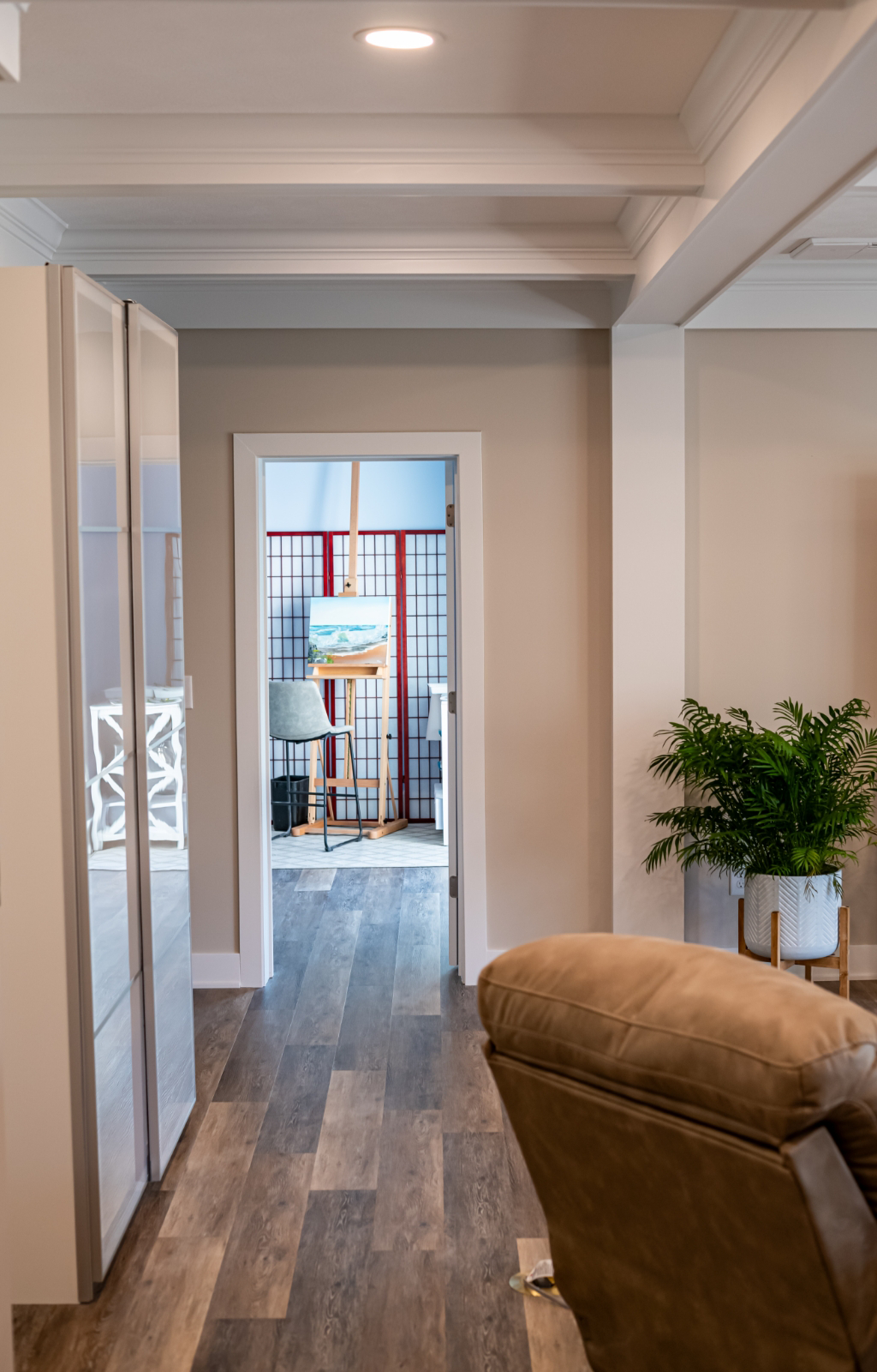 A room with wood flooring, neutral walls, a chair, and a potted plant in the foreground; an open doorway leads to an art studio with easel and art supplies—part of an award-winning home remodel & additions design.