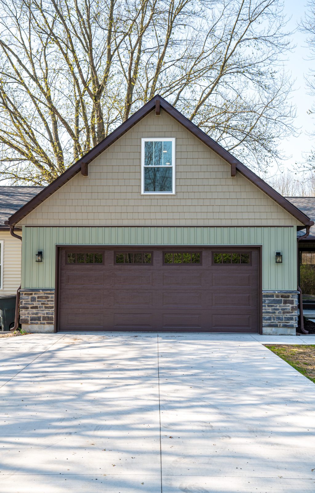 An award-winning home remodel features a two-car garage with light green siding, a brown door, stone accents at the base, and a paved driveway. Leafless trees are visible in the background.