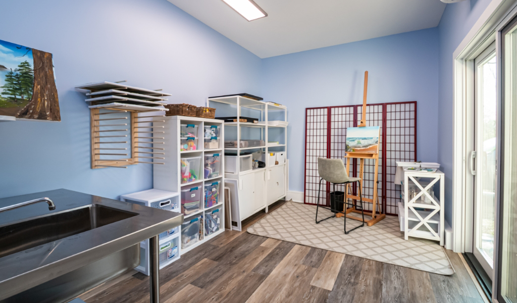A well-organized art studio, part of an award-winning home remodel & additions, with storage shelves, a sink, an easel with a painting, a chair, and large sliding glass doors letting in natural light.