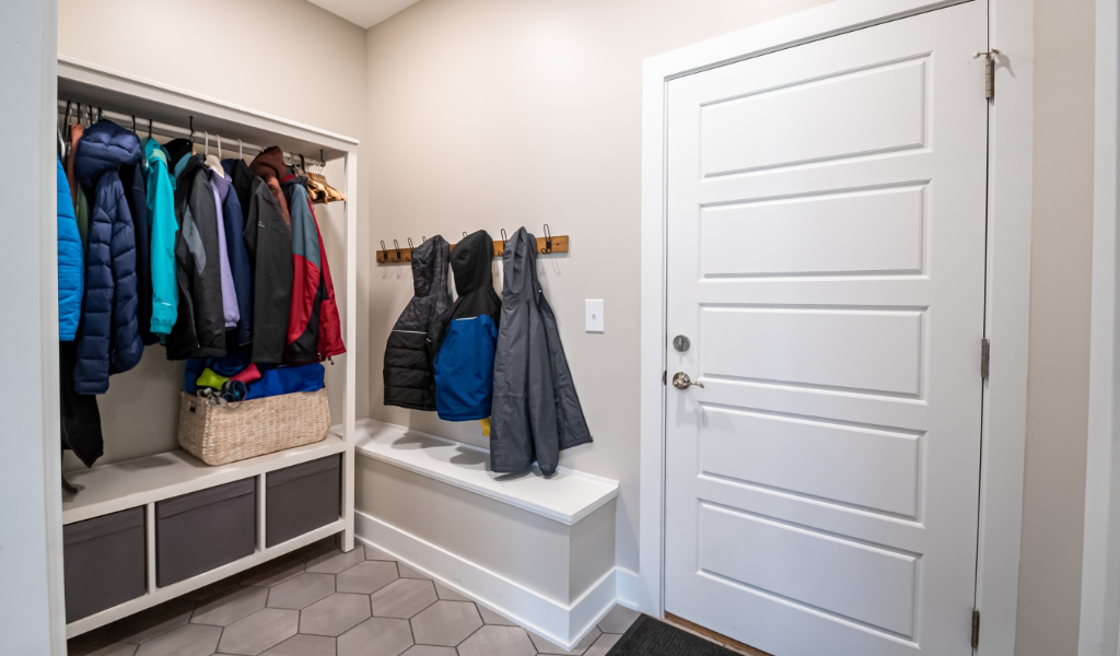 A mudroom from an award-winning home remodel & additions project, featuring a white door, bench, coat hooks, storage basket, and several jackets and coats hanging on hooks and a rod.