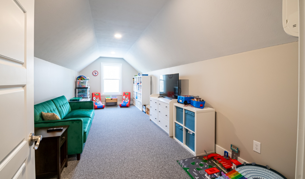 A long, narrow playroom with a green couch, children’s chairs, storage units, toys, and a TV—part of an award-winning home remodel & additions project—illuminated by a window and ceiling lights.