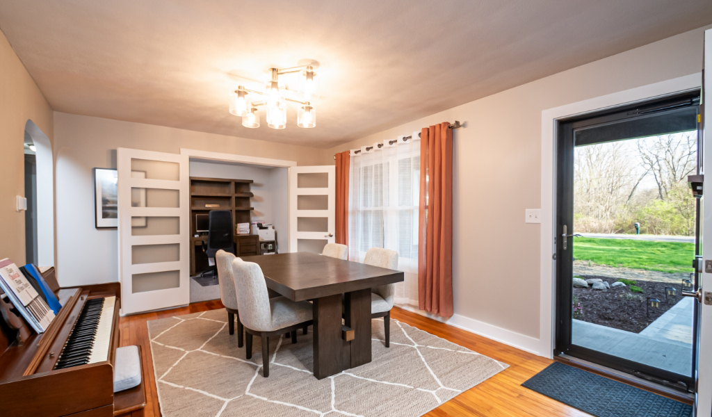 Dining room from an award-winning home remodel & additions, featuring a dark wood table, four upholstered chairs, a piano on the left, glass double doors to an office, large window with curtains, and a glass door leading outside.