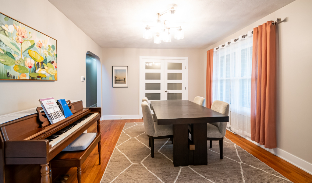 A dining room with a dark wood table, six upholstered chairs, a rug, and a piano with sheet music showcases this award-winning home remodel & additions. Large windows with pink curtains and artwork on the walls complete the inviting space.